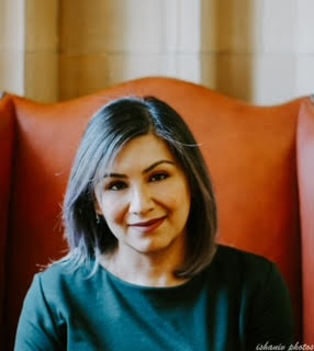 A woman with shoulder-length, straight gray hair sits in an orange chair, smiling slightly. She wears a green long-sleeve shirt.